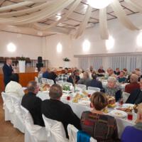 Bal seniora - przy stołach na których znajduje się poczęstunek i napoje siedzą seniorzy - sala jest elegancko przystrojona, niektórzy tańczą