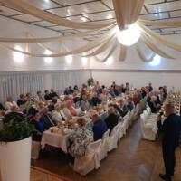 Bal seniora - przy stołach na których znajduje się poczęstunek i napoje siedzą seniorzy - sala jest elegancko przystrojona, niektórzy tańczą