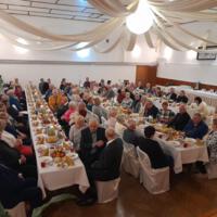 Bal seniora - przy stołach na których znajduje się poczęstunek i napoje siedzą seniorzy - sala jest elegancko przystrojona, niektórzy tańczą