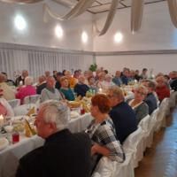 Bal seniora - przy stołach na których znajduje się poczęstunek i napoje siedzą seniorzy - sala jest elegancko przystrojona, niektórzy tańczą