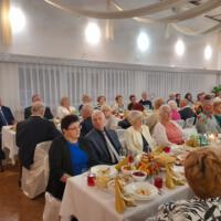 Bal seniora - przy stołach na których znajduje się poczęstunek i napoje siedzą seniorzy - sala jest elegancko przystrojona, niektórzy tańczą