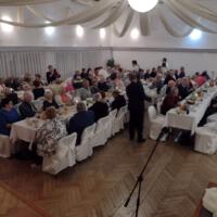 Bal seniora - przy stołach na których znajduje się poczęstunek i napoje siedzą seniorzy - wójt z sołtysem rozdaje pierniki - sala jest elegancko przystrojona