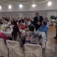 Bal seniora - przy stołach na których znajduje się poczęstunek i napoje siedzą seniorzy - wójt z sołtysem rozdaje pierniki - sala jest elegancko przystrojona
