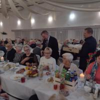 Bal seniora - przy stołach na których znajduje się poczęstunek i napoje siedzą seniorzy - wójt z sołtysem rozdaje pierniki - sala jest elegancko przystrojona