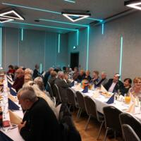 Przy stołach siedzi kilkadziesiąt osób w starszym wieku. Na stołach napoje zastawa itp. Sala jest podświetlona na niebiesko