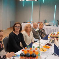 Przy stołach siedzi kilkadziesiąt osób w starszym wieku. Na stołach napoje zastawa itp. Sala jest podświetlona na niebiesko