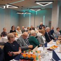 Przy stołach siedzi kilkadziesiąt osób w starszym wieku. Na stołach napoje zastawa itp. Sala jest podświetlona na niebiesko