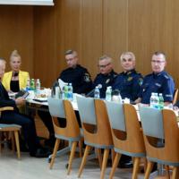 W sali przy stole siedzi kilka osób, niektóre osoby w niebieskich mundurach, na stołach napoje, czajniki, filiżanki i mikrofony
