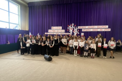 Uczestnicy konkursu ustawieni w rzędzie do wspólnego zdjęcia - wśród nich wójt, dyrektor szkoły i radny Andrzej Tyrkiel, sala gimnastyczna udekorowana fioletowymi zasłonami oraz napisami RADOSNA NIEPODLEGŁOŚCI gminny konkurs pieśni patriotycznych oraz Woln