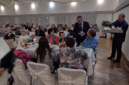 Bal seniora - przy stołach na których znajduje się poczęstunek i napoje siedzą seniorzy - wójt z sołtysem rozdaje pierniki - sala jest elegancko przystrojona