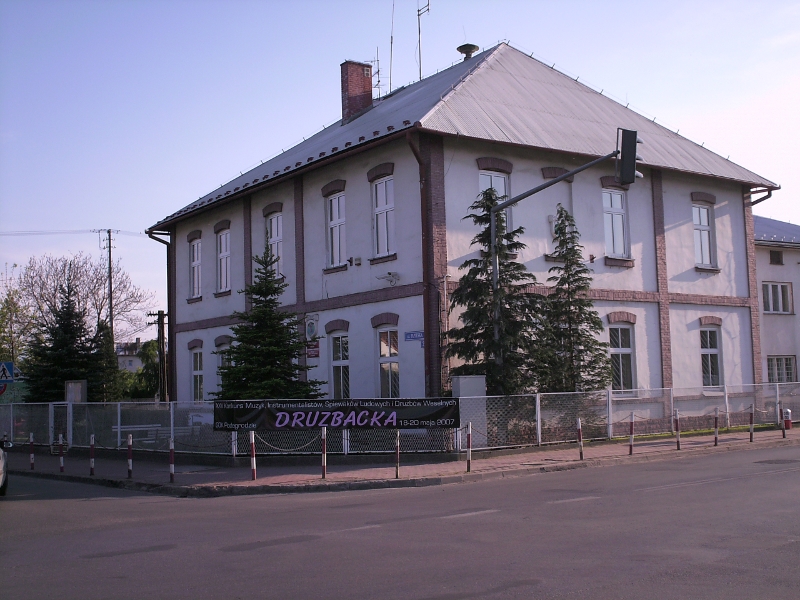 Budynek urzędu gminy jednopiętrowy ze spadzistym dachem - widok ze skrzyżowania, przed budynkiem choinki, ogrodzenie, chodnik i część skrzyżowania