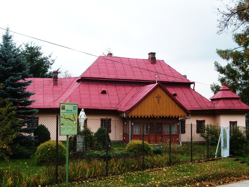 Niewielki dworek kryty czerwoną blachą, budynek ogrodzony, wejście do budynku drewniane, przed budynkiem tablica informacyjna oraz niewielki ogród